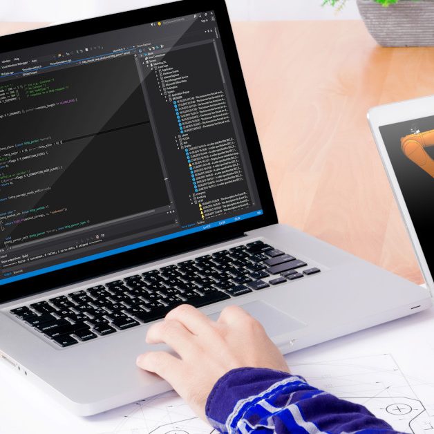 Software engineer's workplace. Close up of engineer's hands working on the notebook and holding tablet with kuka on the display. Note: Program code was got from github with free license for commercial use (MIT)